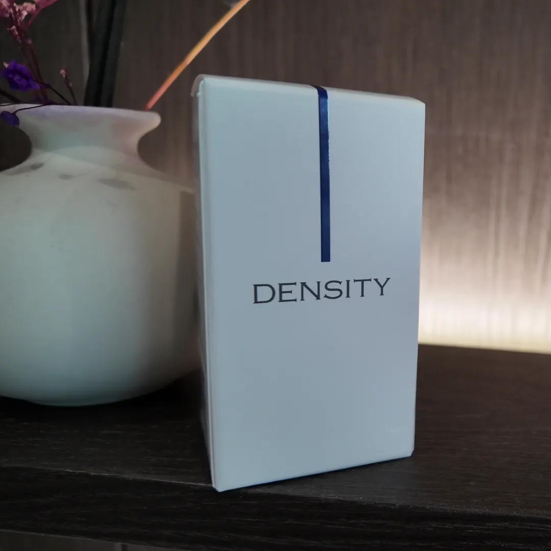 어두운 선반 위 흰 꽃병 옆에 DENSITY라고 인쇄된 흰색 제품 상자가 세워져 있는 모습