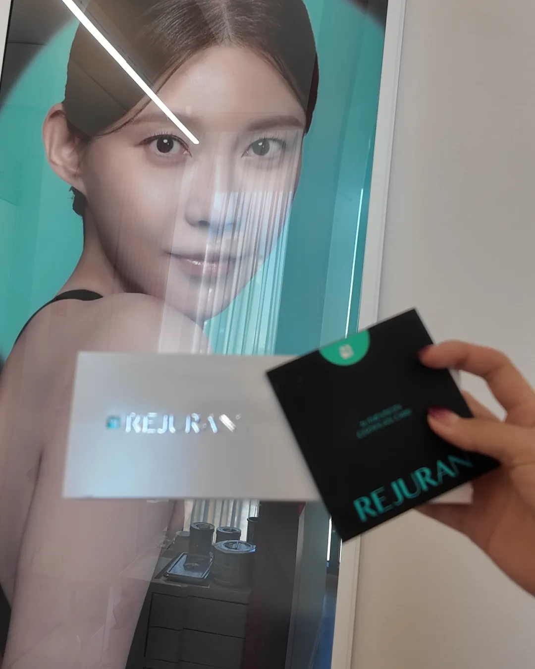 REJURANの壁面ポスターの前で、REJURANの箱と認証カードを持つ手
