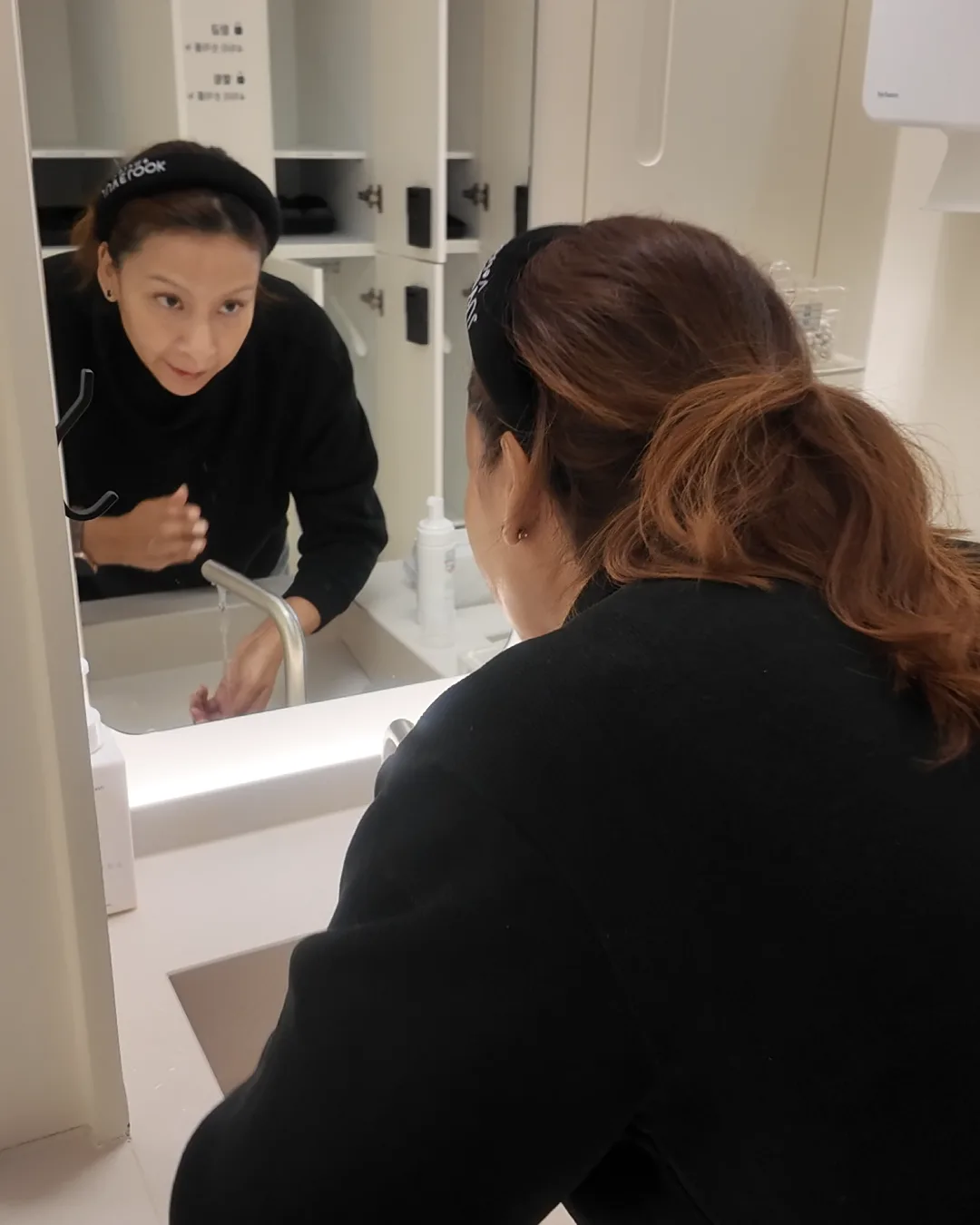 黒いトップスの女性が洗面台に身をかがめて顔をすすぎ、その姿が鏡に映っている様子