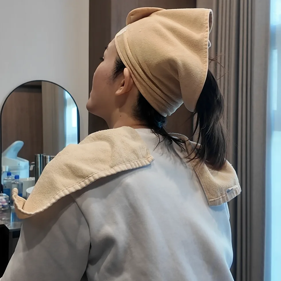 A person seen from behind, seated in a treatment room with a towel around the head and shoulders
