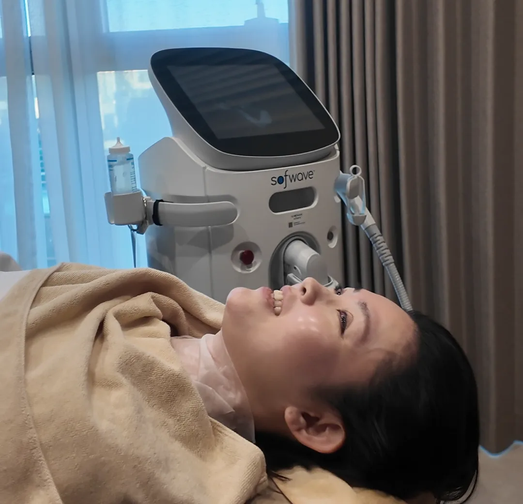 A person lying on a treatment chair in a bright room with a Sofwave console beside the window